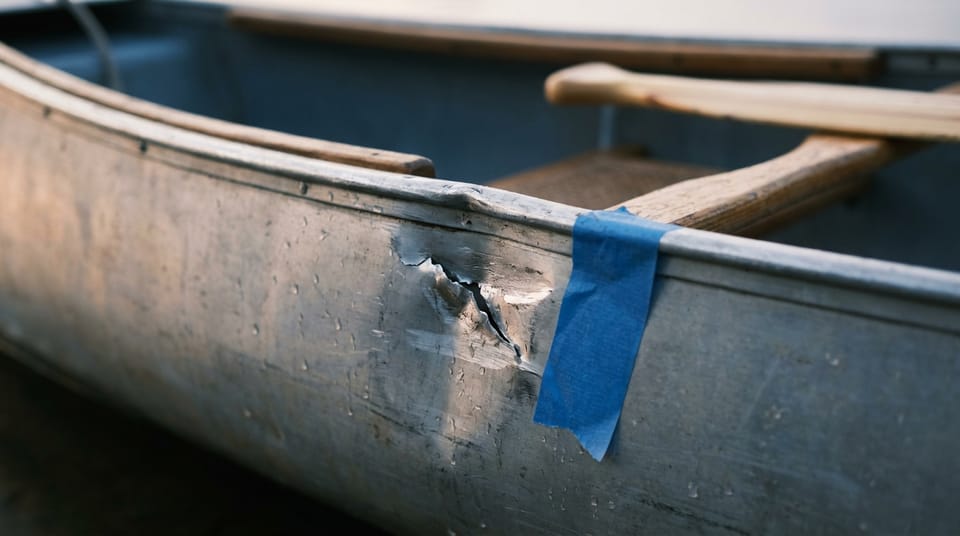 Close view of a fresh dent and scratch in the aluminum side of a canoe, a strip of blue painter's tape stuck beside it, water droplets on the metal.