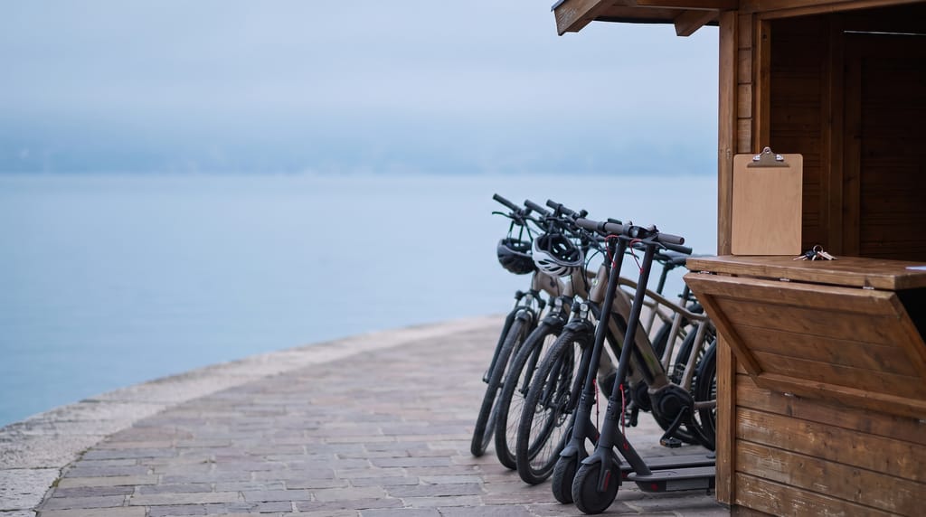 Electric bikes and two electric kick scooters parked beside a small wooden rental stand on a lakeside promenade in morning mist, helmets on the handlebars and a blank clipboard and keys on the counter.