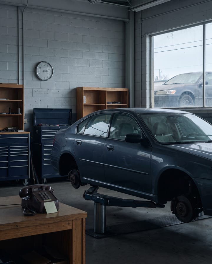 A plain gray sedan up on a lift with its front wheels off in a service bay, an old desk phone with a blank sticky note on the workbench, a wall clock, and a second dusty car through the window.
