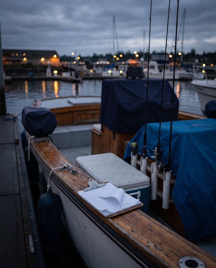 A charter boat's cockpit at the dock before dawn: rods in their holders, a cooler, a waterlogged clipboard of paper waivers curling on the gunwale, harbor lights beyond.